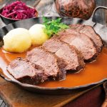 Traditional braised marinated German Sauerbraten from beef with potato dumplings and blue kraut in spicy brown sauce as closeup in a rustic wrought iron pan