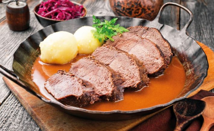 Traditional braised marinated German Sauerbraten from beef with potato dumplings and blue kraut in spicy brown sauce as closeup in a rustic wrought iron pan